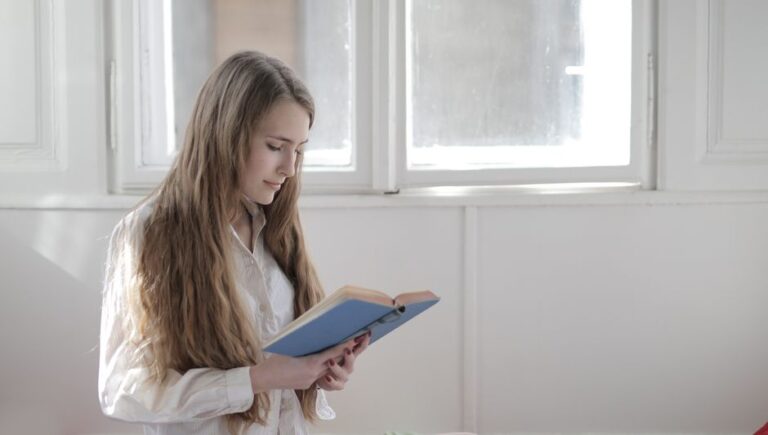 A young woman engrossed in a book while standing near a sunlit window indoors.