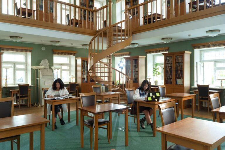 Two women studying in a quiet, well-lit library with spiral stairs and classic decor.